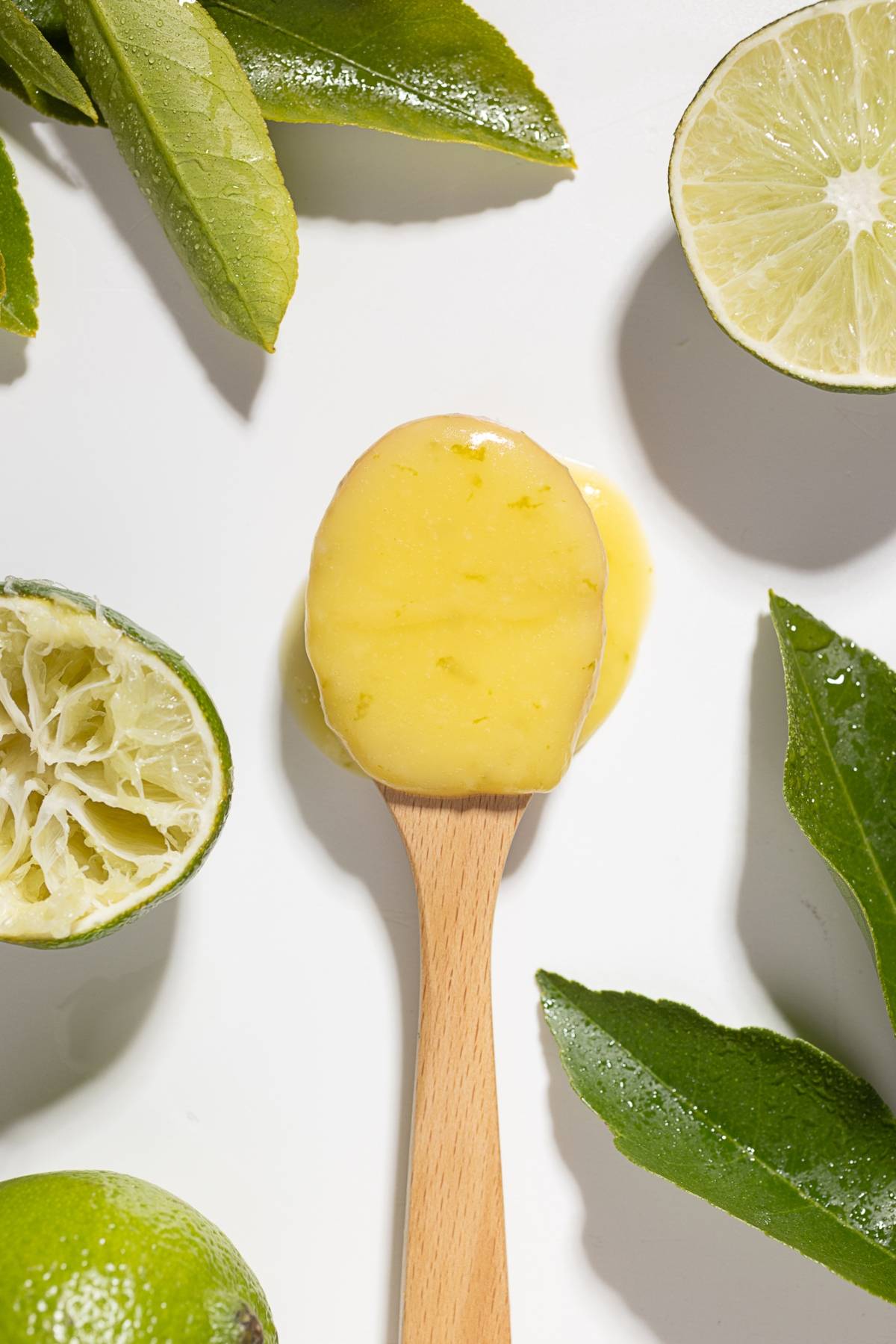 A wooden spoon with lemon curd, surrounded by lime halves and green leaves on a white background.