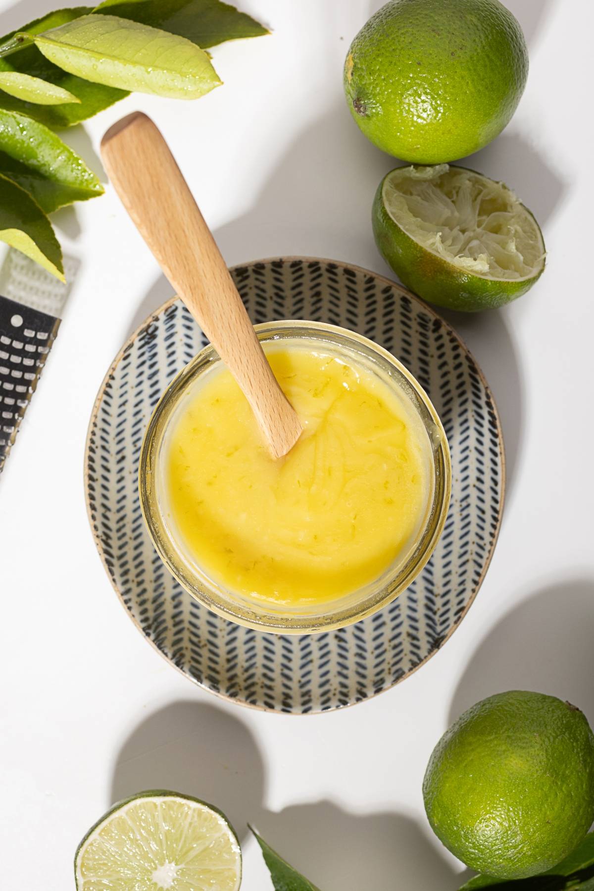 A bowl of yellow lime curd with a wooden spoon, surrounded by fresh and cut limes on a patterned plate.