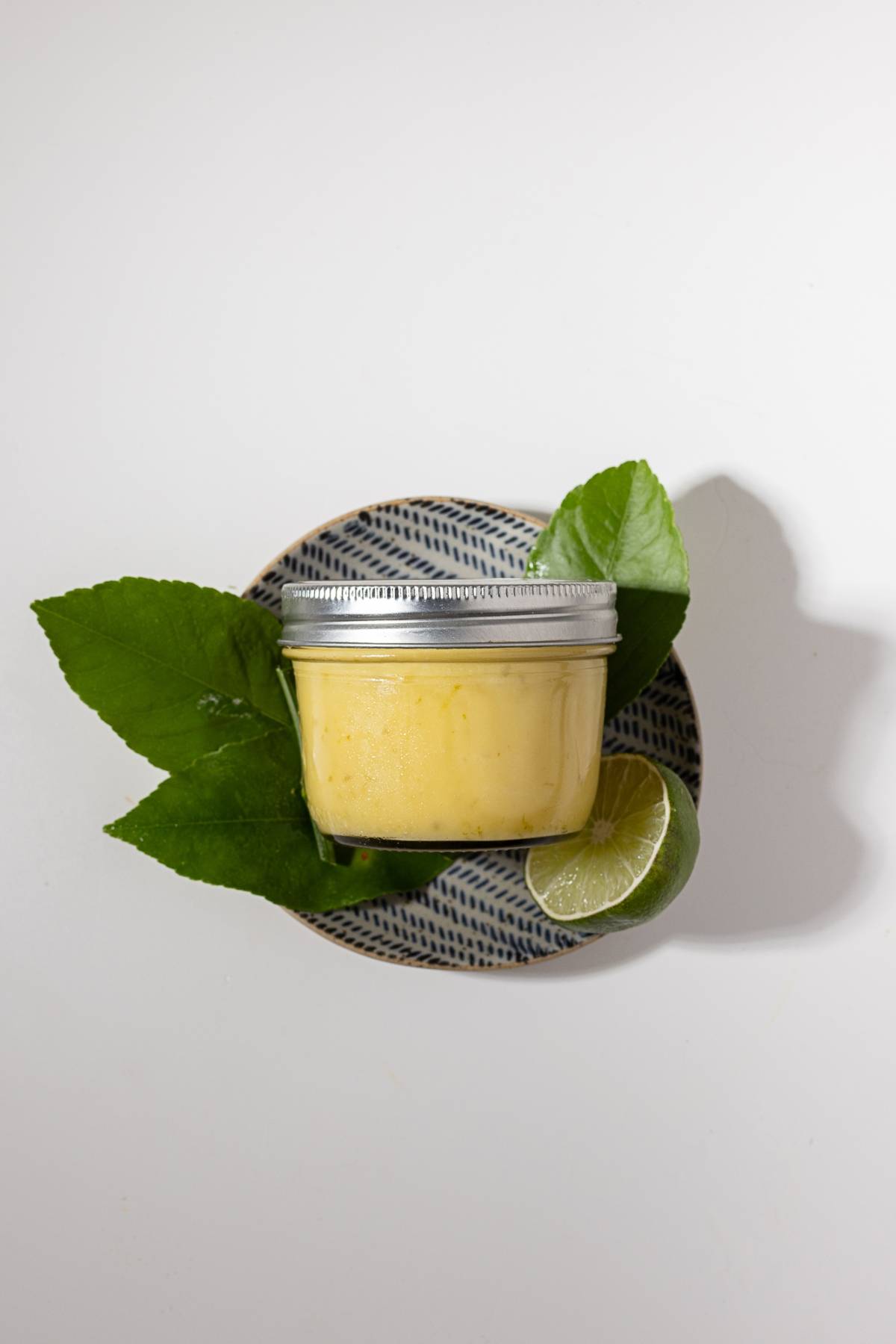 A jar of yellow cream on a patterned plate with green leaves and a sliced lime.