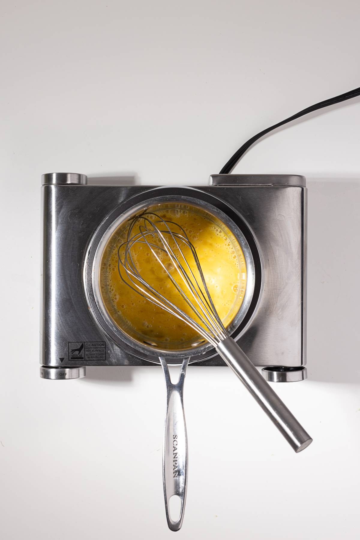 A whisk in a bowl of beaten eggs on a metal hot plate, viewed from above.