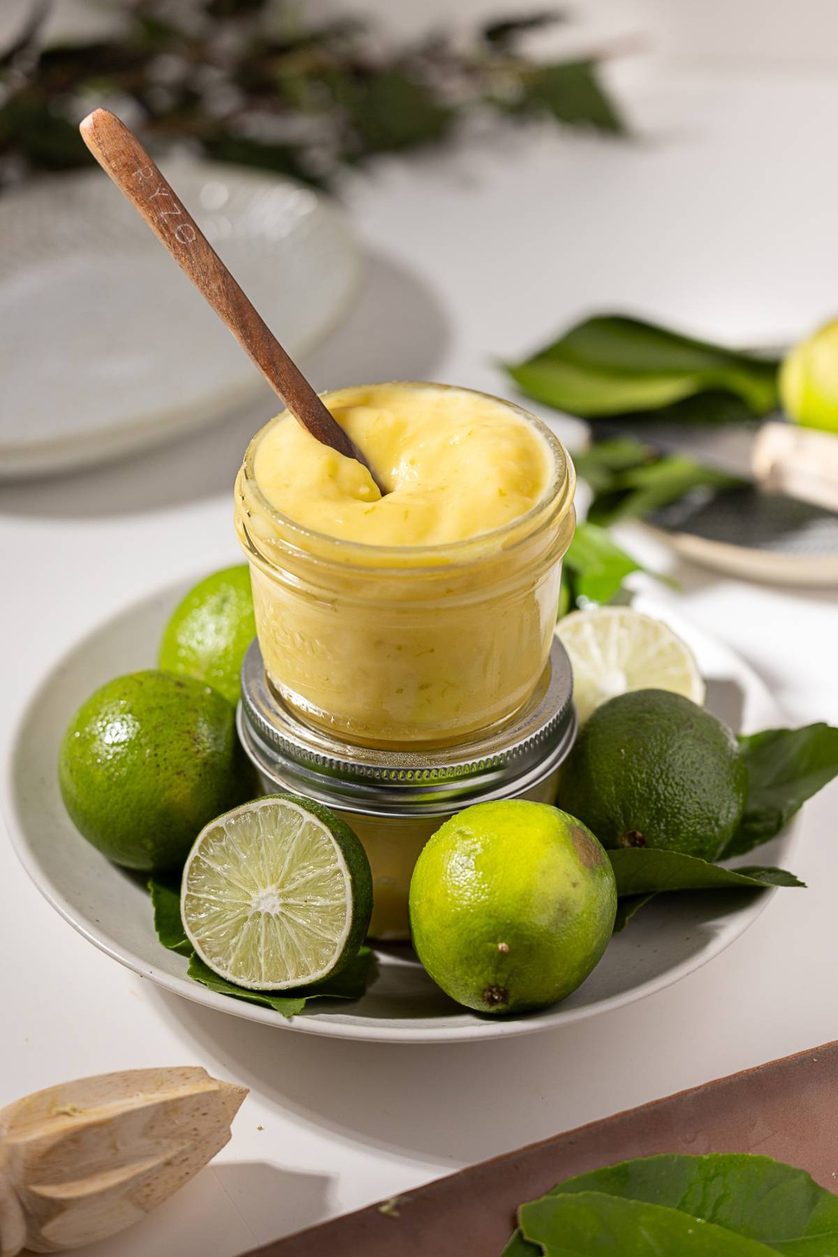 A jar of creamy yellow curd with a spoon, surrounded by whole and sliced limes on a plate.
