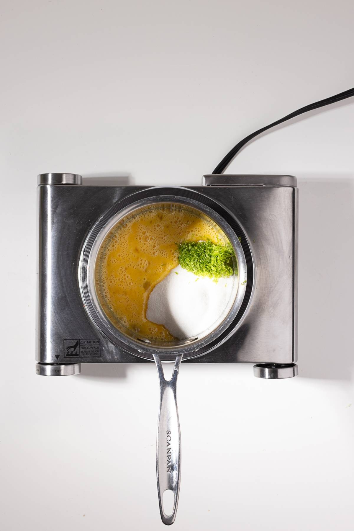 A saucepan with whisked eggs, sugar, and lime zest sits on a silver electric stove against a white background.