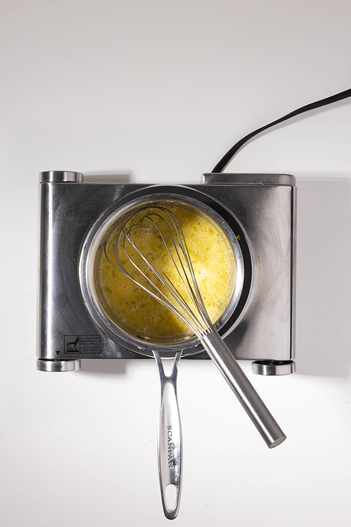 Metal whisk in a saucepan with beaten eggs on an electric stovetop, viewed from above.