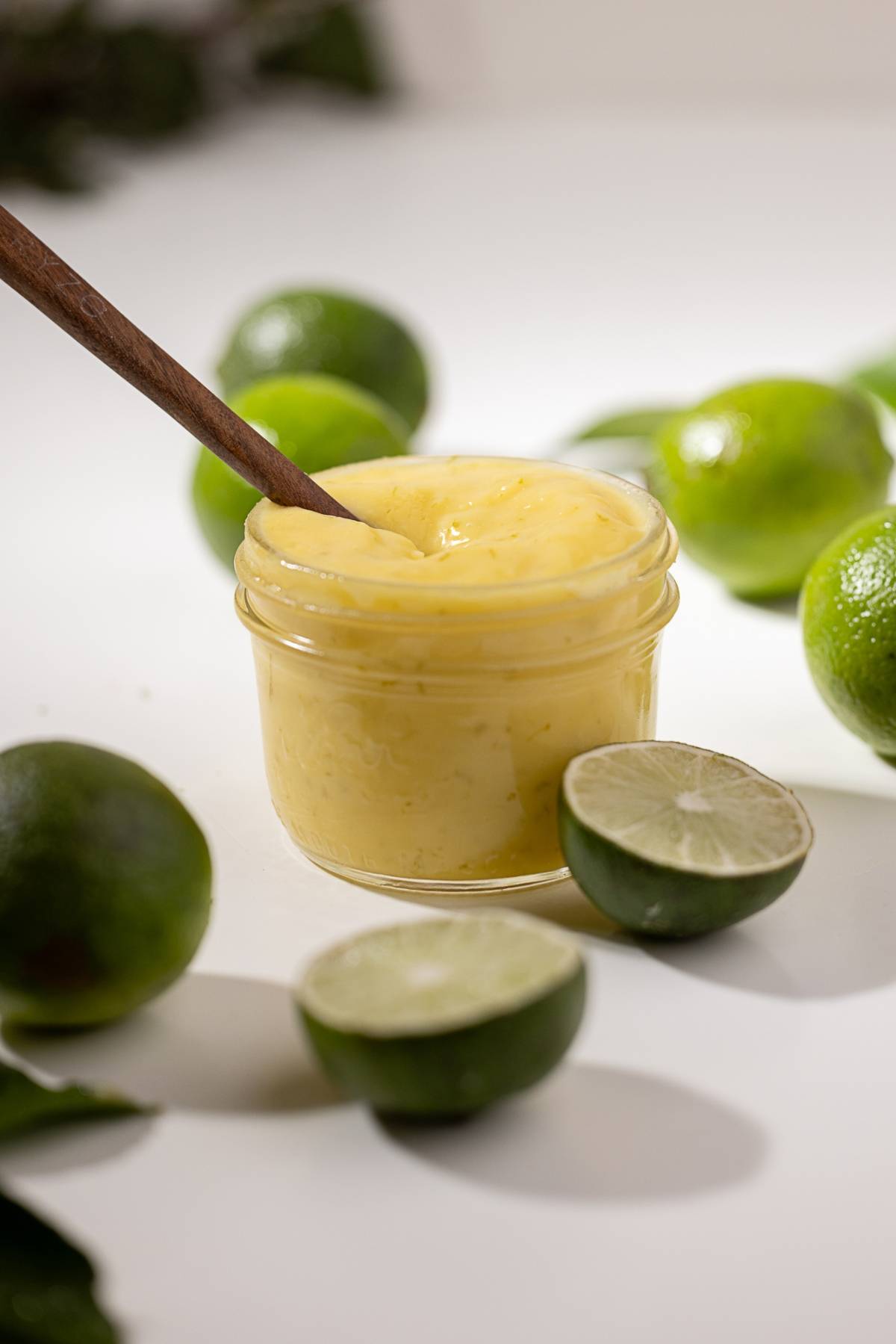A glass jar of creamy lime curd with a wooden spoon, surrounded by fresh whole and halved limes.