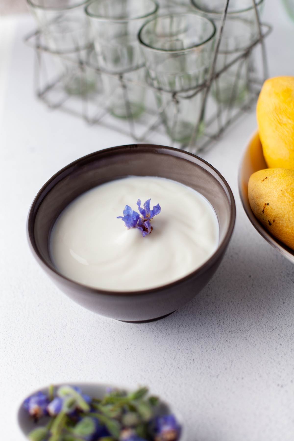 A bowl of yogurt topped with a purple flower sits beside a bowl of yellow fruits on a white surface, making for an inviting mango yogurt dessert and a healthy dessert for kids.