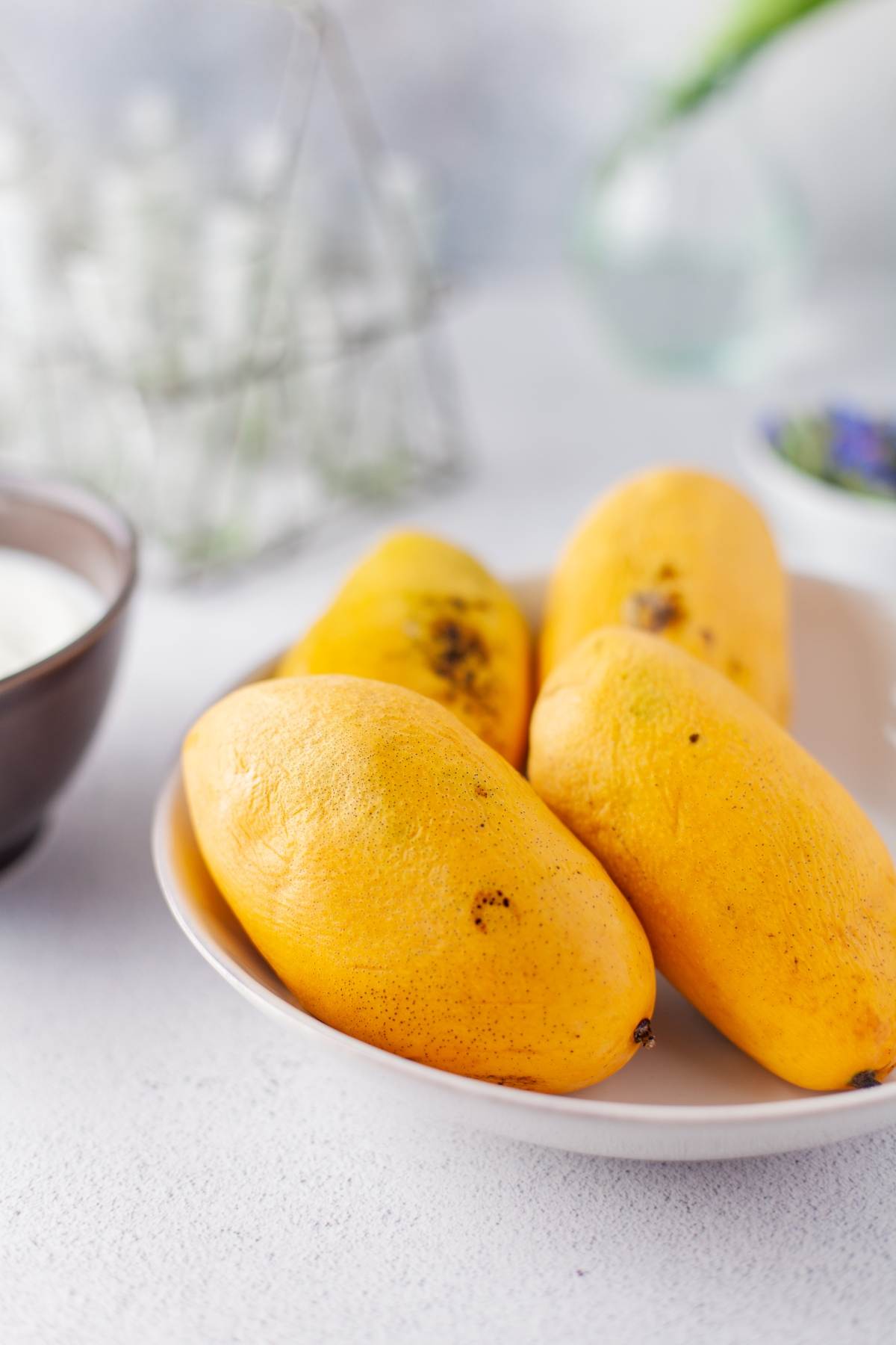 Four ripe yellow mangoes on a white plate, with a blurred background and a bowl on the side—perfect for creating a refreshing Mango Yogurt Dessert or trying your favorite healthy mango mousse recipe.