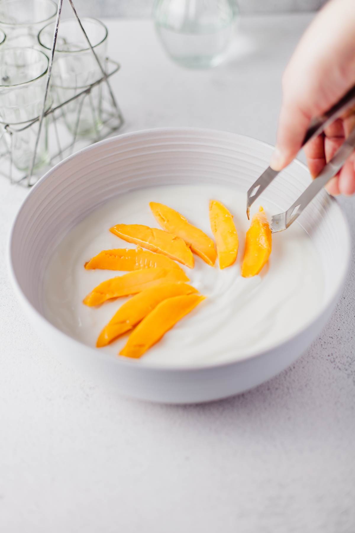 A hand places mango slices on yogurt in a white bowl using metal tongs, creating a colorful Mango Yogurt Dessert—an easy, healthy dessert for kids.