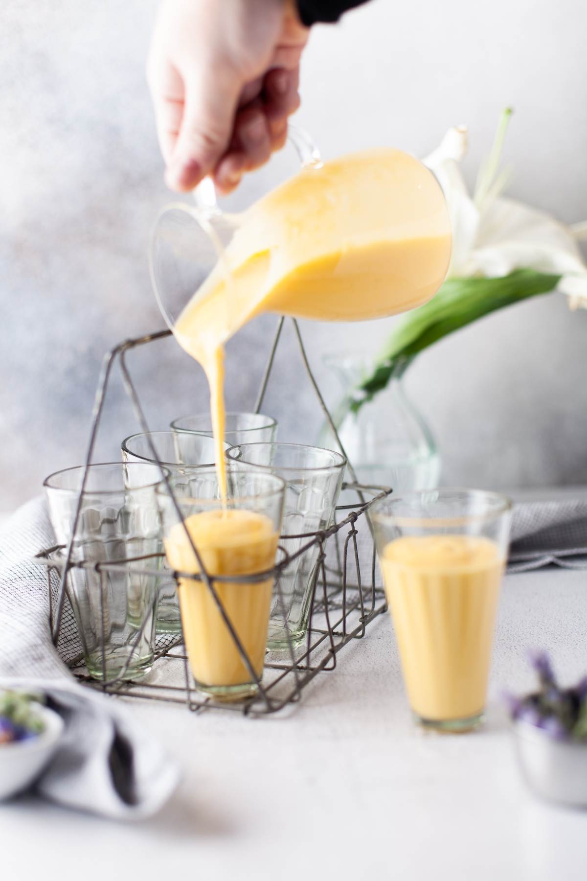 A hand pours creamy Mango Yogurt Dessert lassi from a pitcher into a glass, with other empty glasses nearby—an irresistible and healthy dessert for kids.