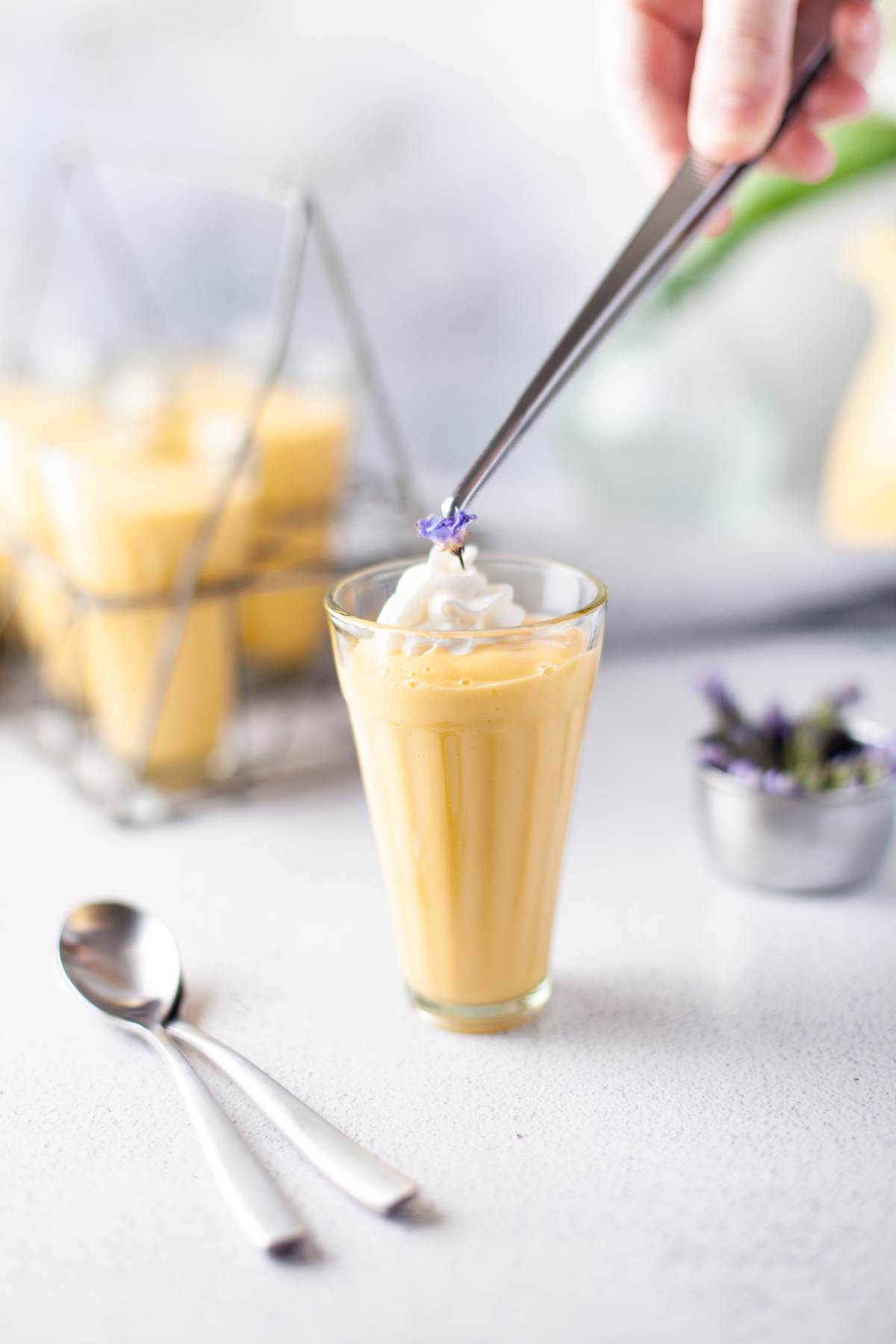 A hand adds a purple flower to whipped cream atop a glass of yellow mango yogurt dessert; two spoons are nearby.
