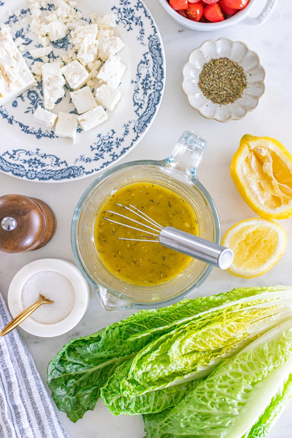 Salad ingredients with feta, cherry tomatoes, lemon halves, lettuce, seasoning, and vinaigrette in a glass bowl.