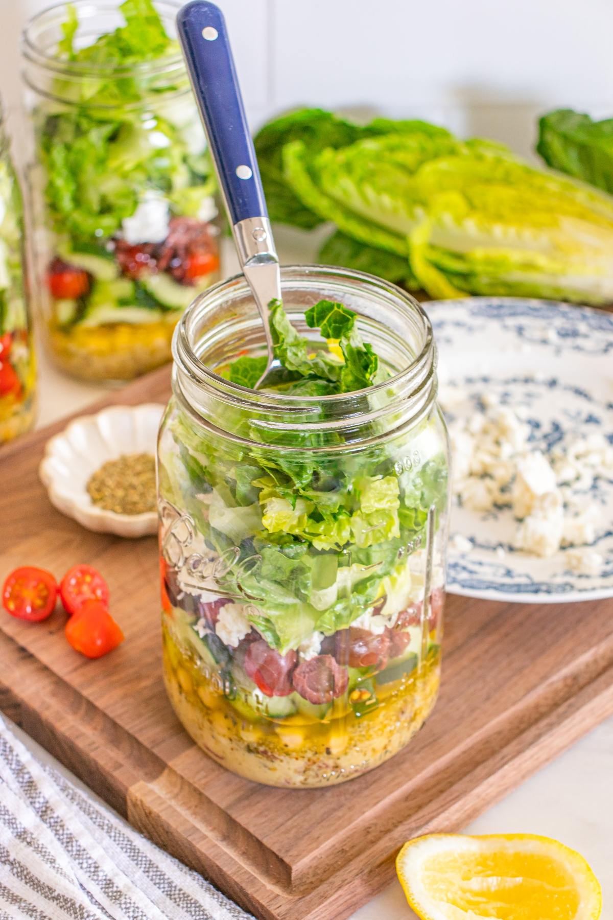 A mason jar layered with salad ingredients and a fork, surrounded by fresh veggies and cheese on a wooden board.