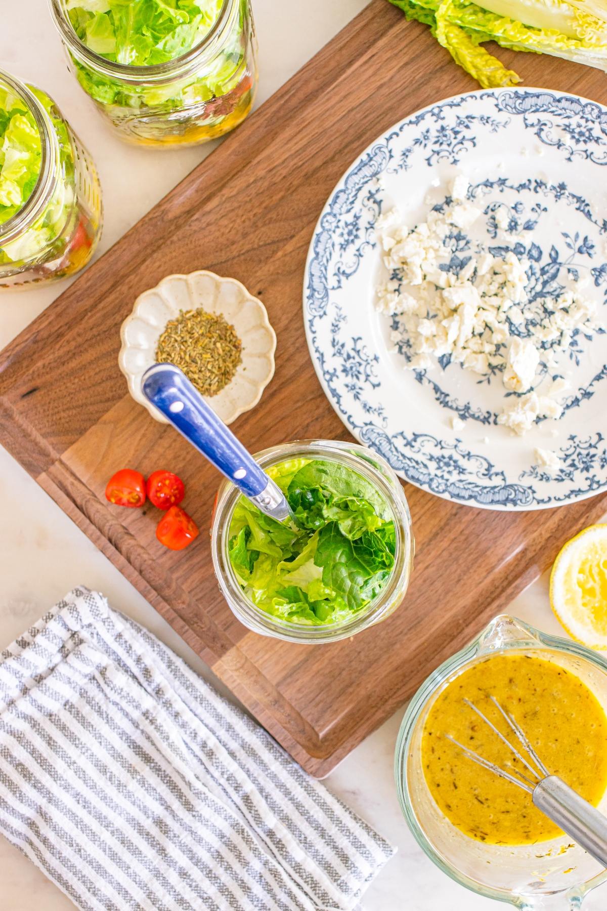 Overhead view of salad jars, blue cheese, tomatoes, herbs, and dressing on a wooden cutting board.