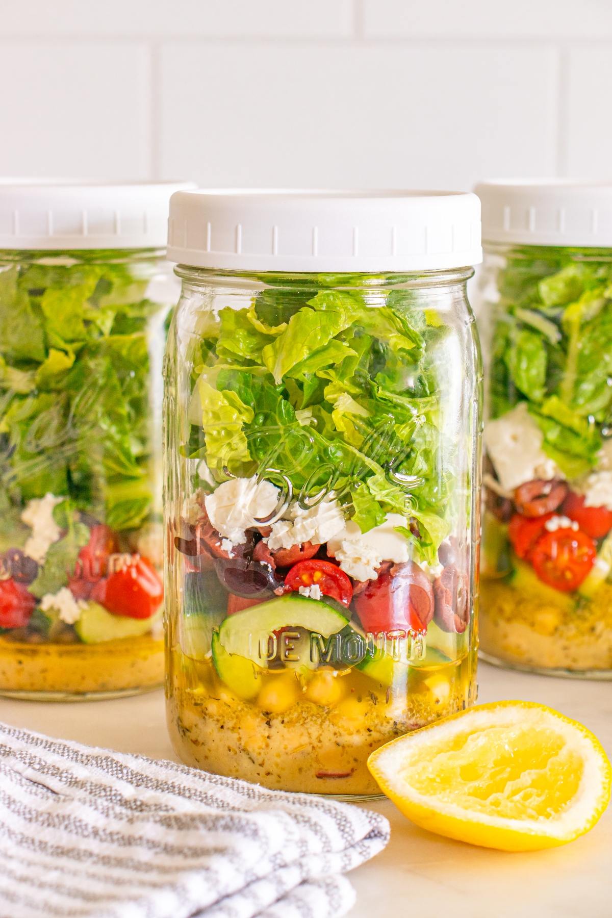 Three mason jars filled with layered salad ingredients, next to a sliced lemon and striped napkin.