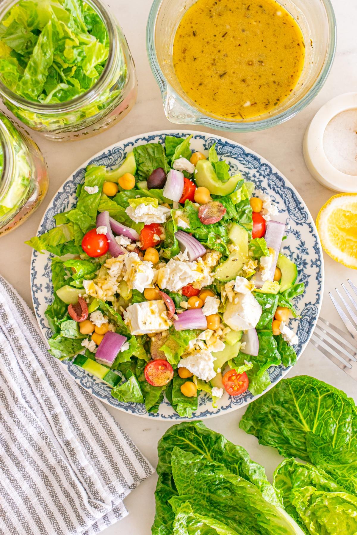 A plate of Greek salad with feta, chickpeas, tomatoes, and onions, next to dressing, lettuce, and lemon.