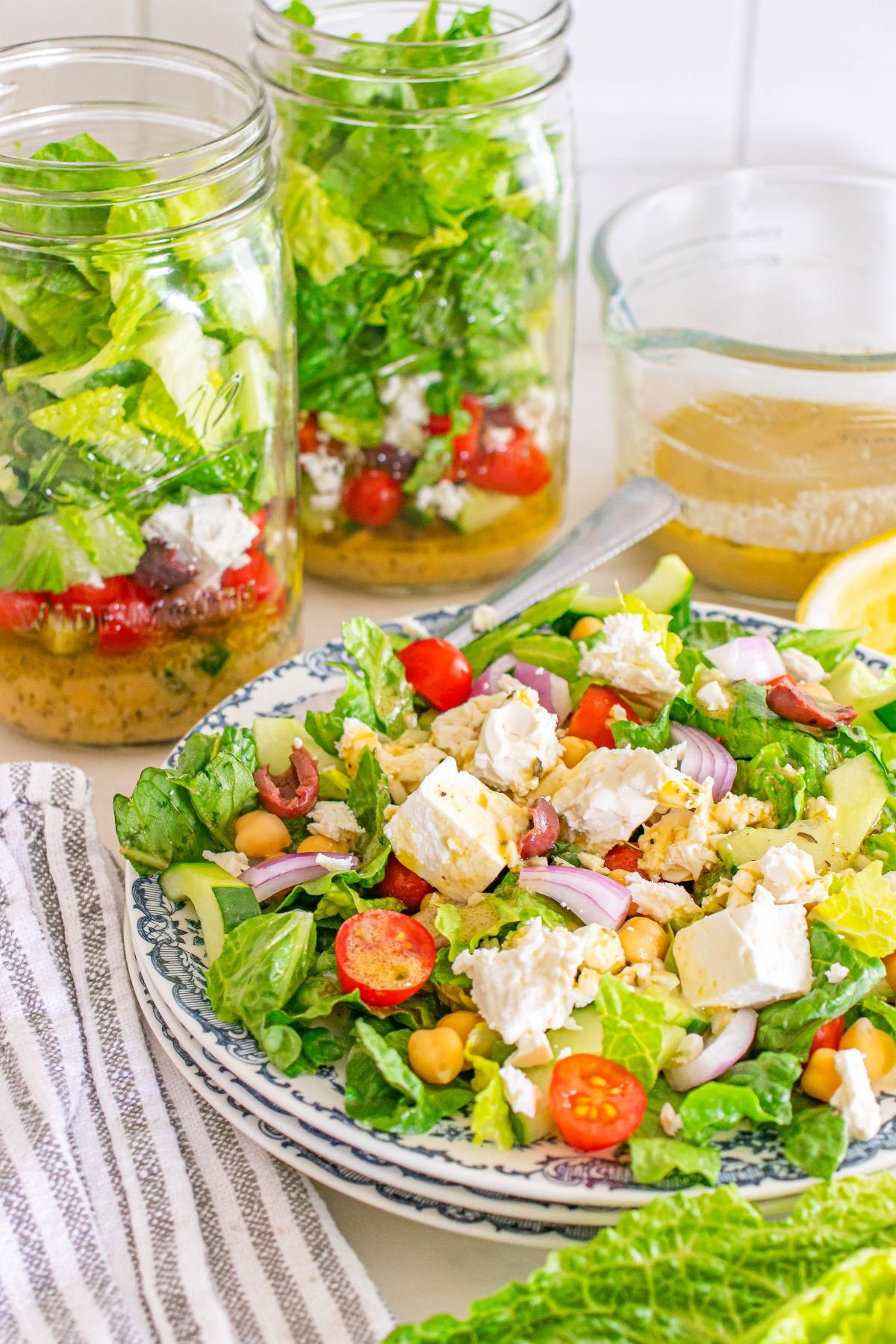 A plate of Greek salad with feta, chickpeas, and veggies, with two salad jars and dressing in the background.