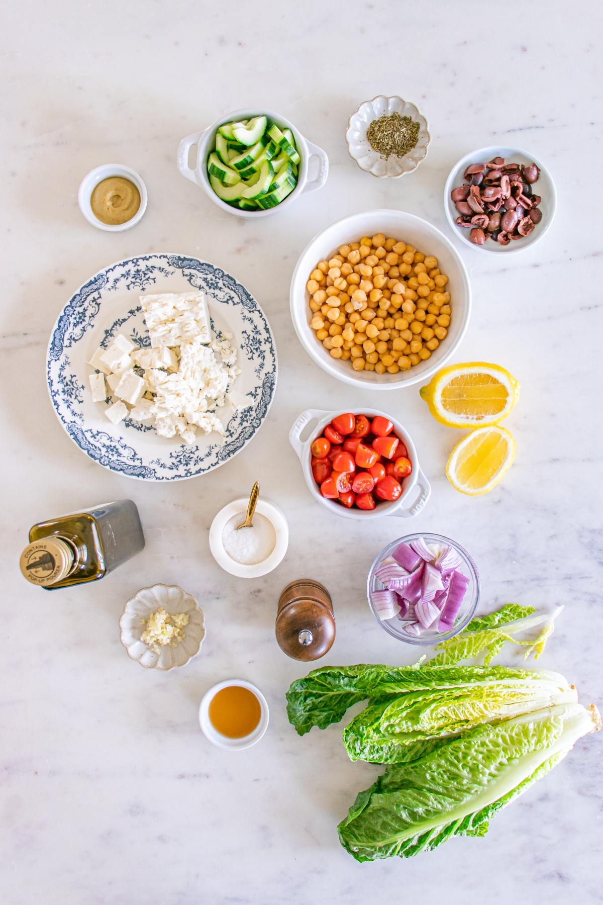 Salad ingredients on a white surface: lettuce, tomatoes, chickpeas, feta, olives, cucumber, onion, lemon, and seasonings.