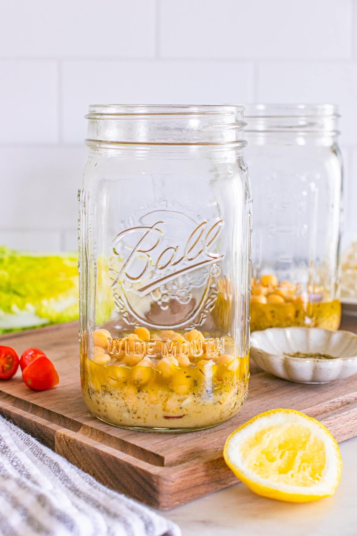 A mason jar with chickpeas and dressing inside sits on a wooden board near a halved lemon and tomatoes.