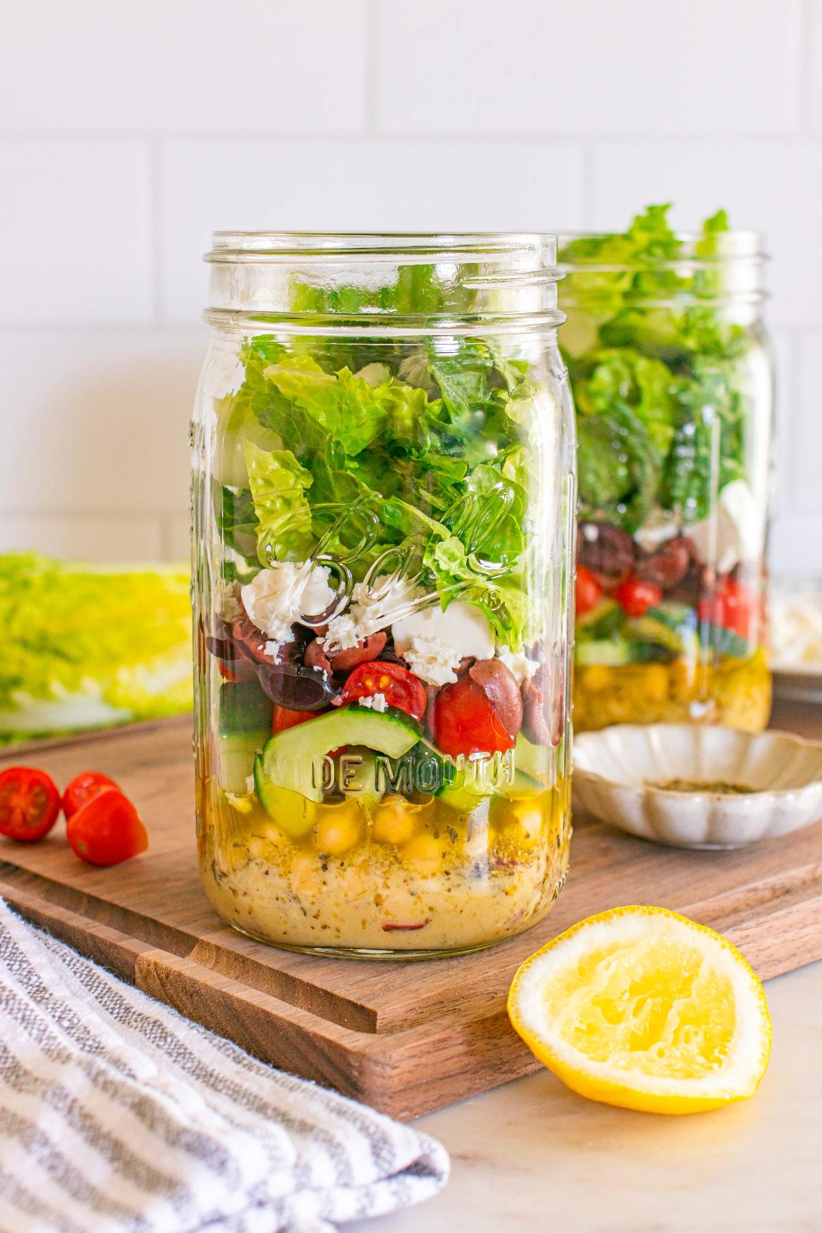 Two mason jars filled with layered salad ingredients, including chickpeas, cucumbers, tomatoes, and lettuce.