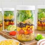 Four mason jars layered with classic Mason Jar Taco Salad ingredients, including beans, corn, tomatoes, lettuce, and shredded cheese.