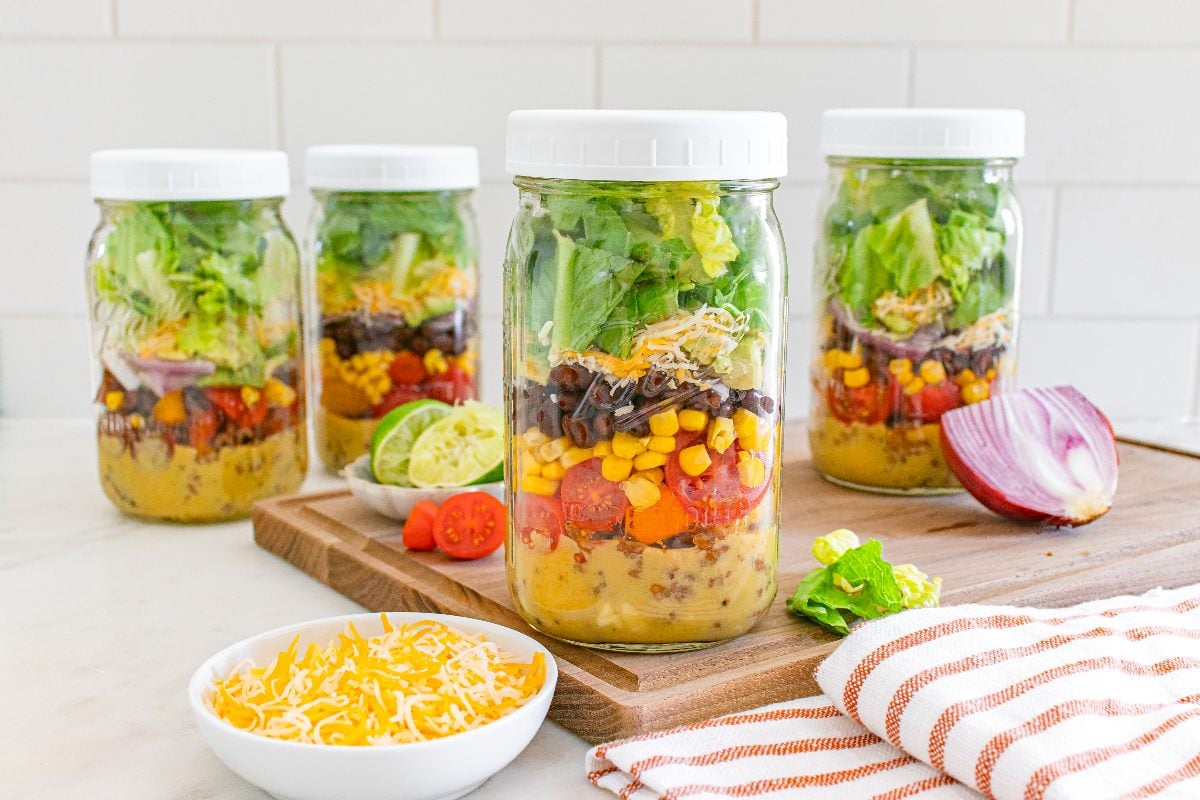 Four mason jars layered with Mason Jar Taco Salad ingredients sit next to veggies, cheese, and a striped towel on a kitchen counter.