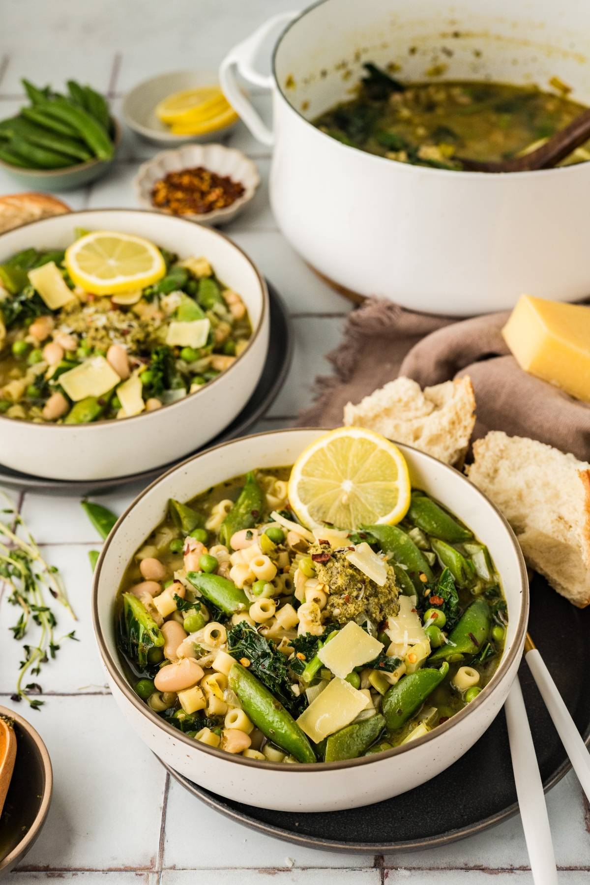 Two bowls of vegetable soup with pasta, beans, and lemon slices, served with bread and a pot in the background.