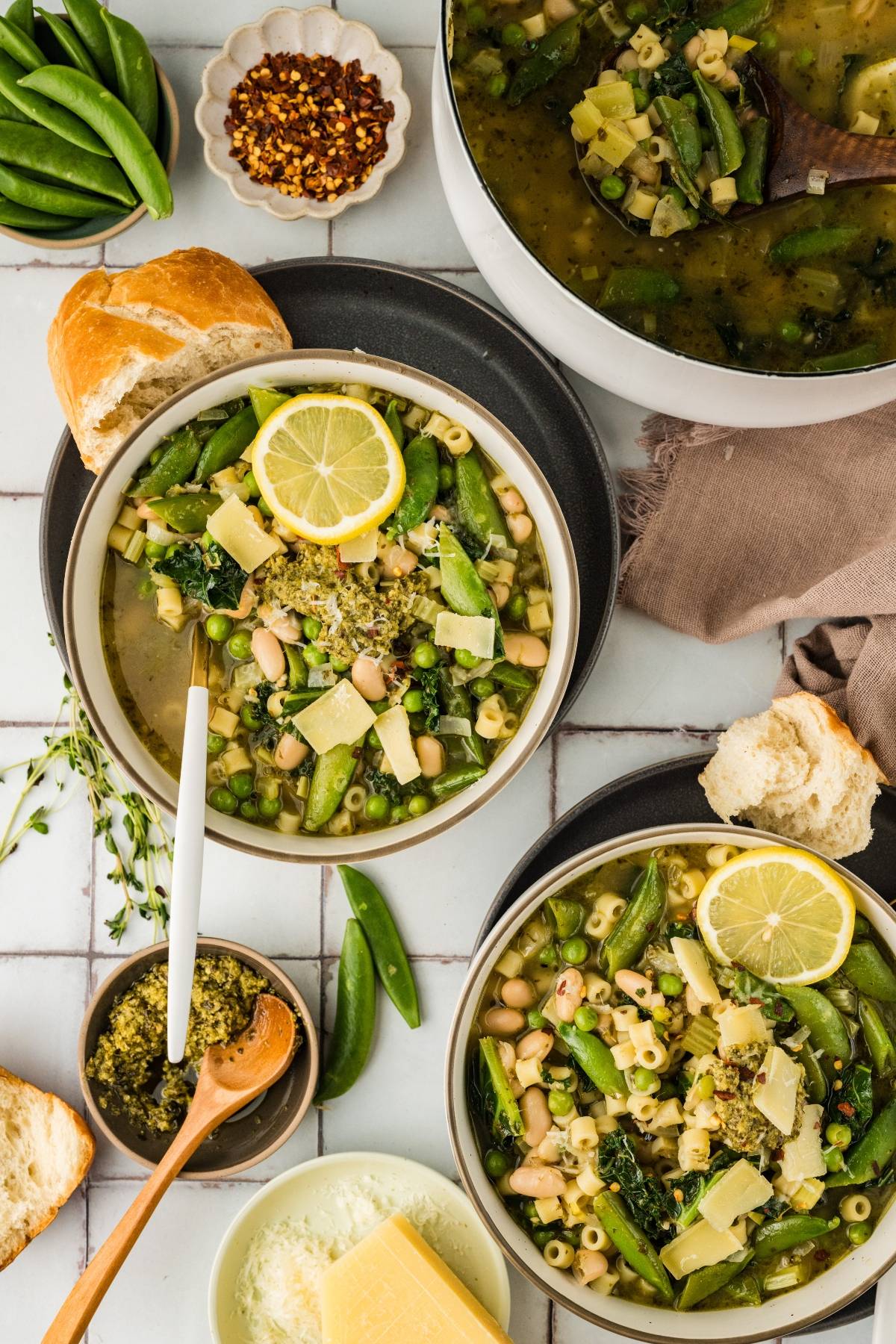 Two bowls of vegetable soup with pasta, beans, lemon slices, and bread on a tiled surface.
