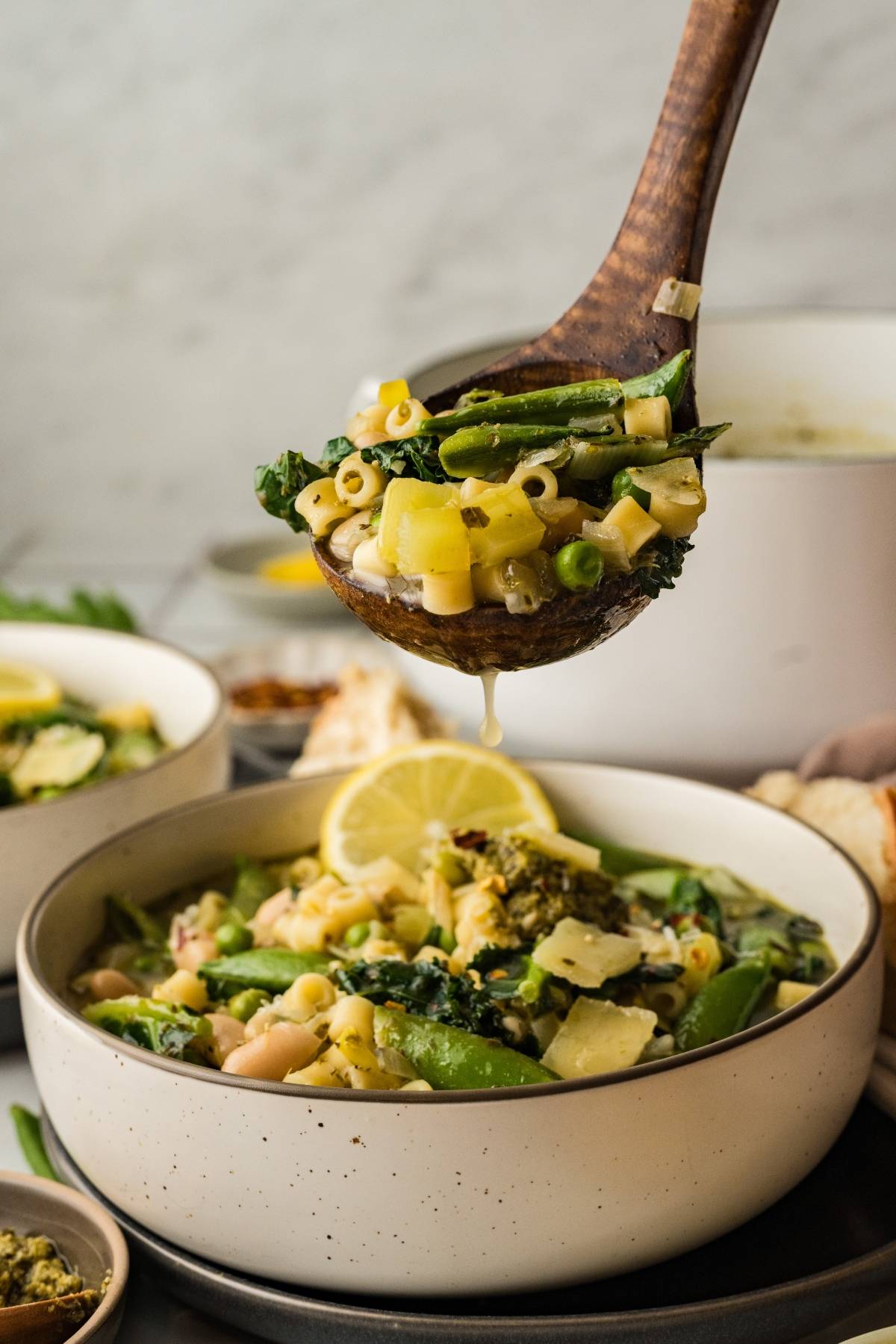 A wooden ladle serving vegetable soup with pasta, green beans, and greens into a white bowl.