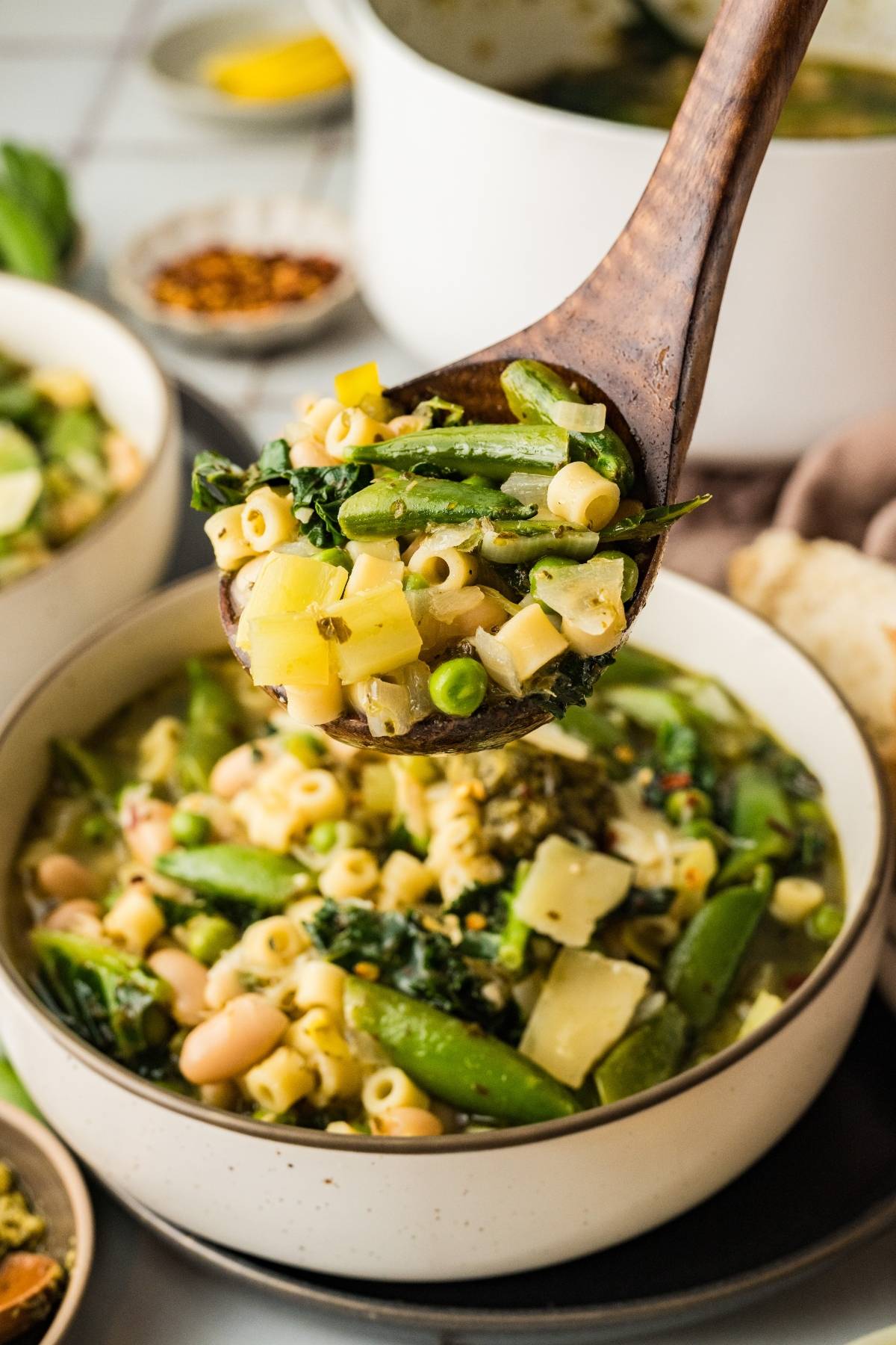 A wooden spoon serves vegetable soup with pasta, beans, and greens over a bowl.