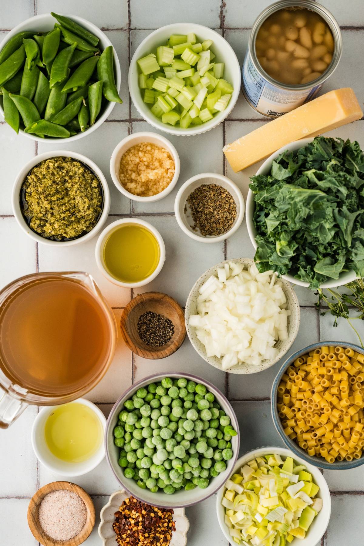 Bowls of vegetables, pasta, seasonings, pesto, broth, and a can of beans arranged on a white tiled surface.