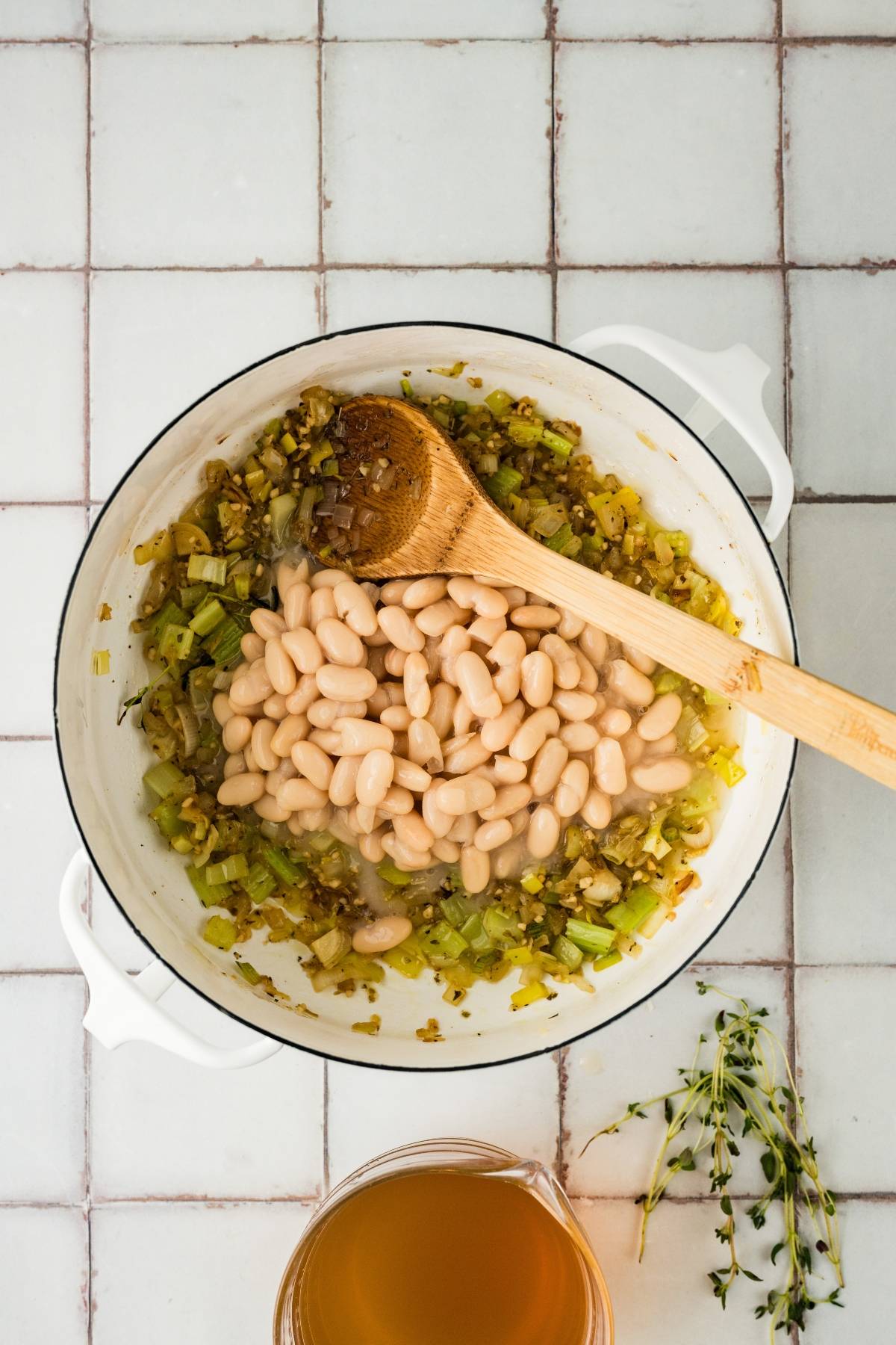 White beans in a pot with sautéed vegetables, wooden spoon, thyme, and broth on a tiled surface.