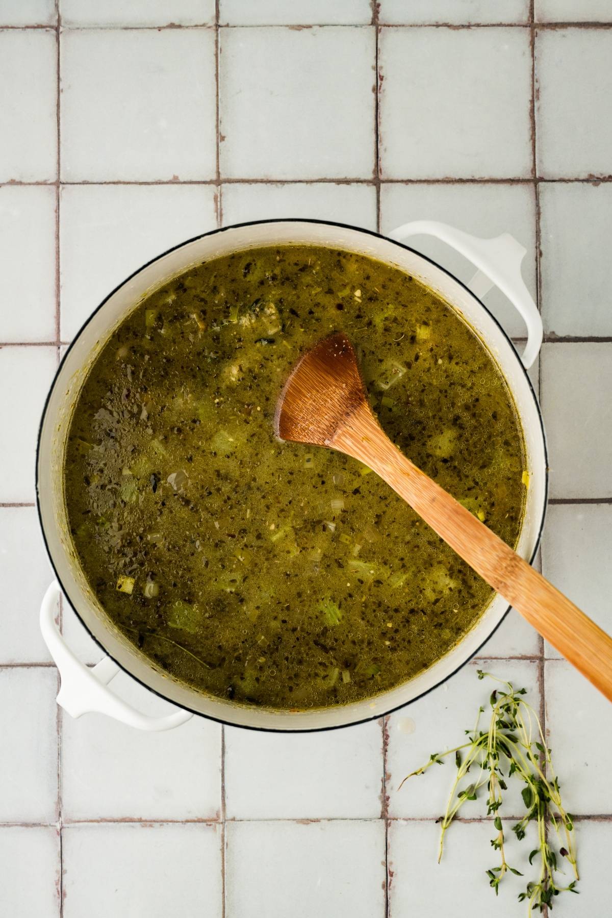 A pot of greenish soup with herbs, stirred by a wooden spoon, sits on a tiled surface with thyme sprigs nearby.