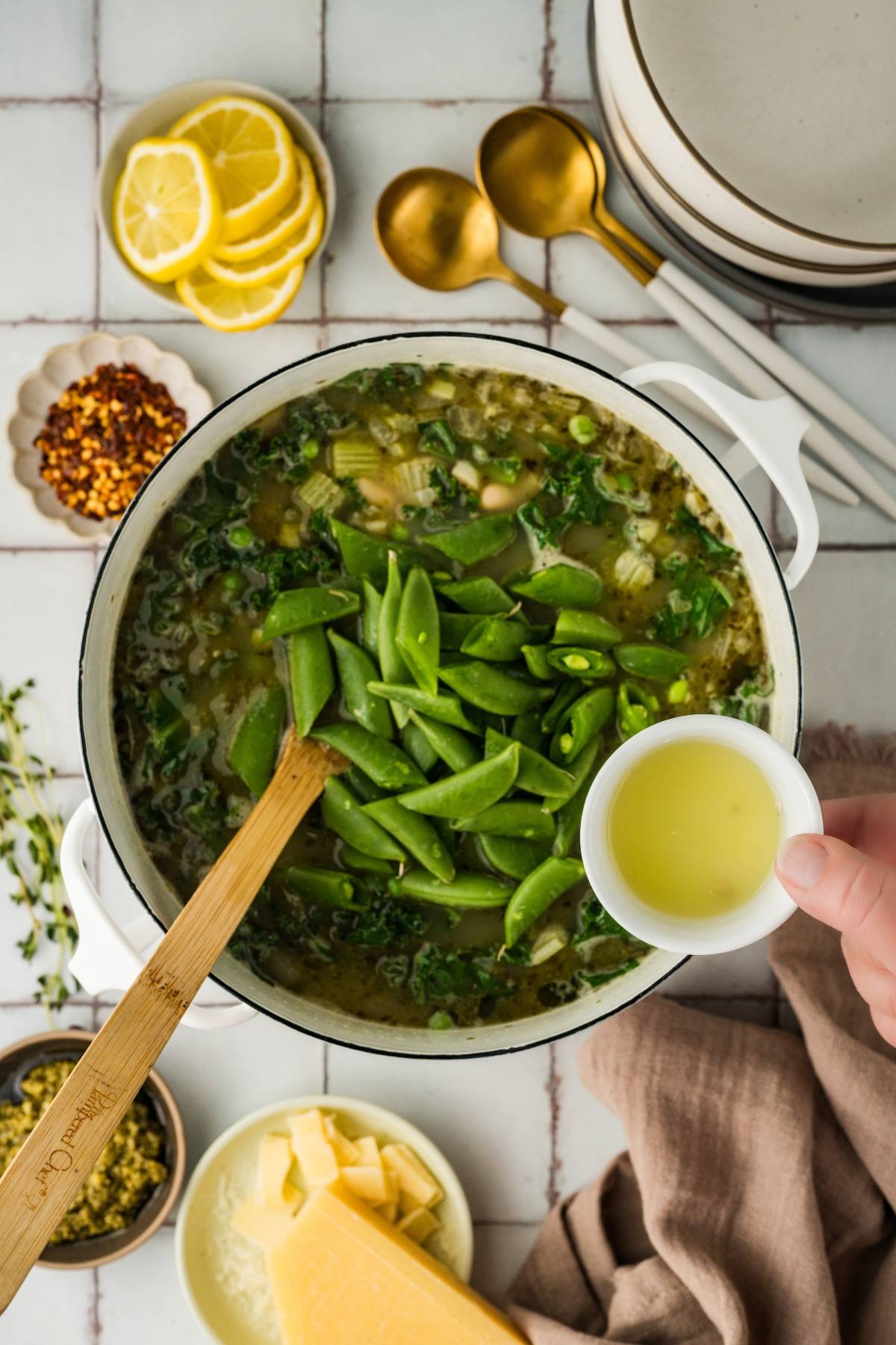 A hand adds lemon juice to a pot of soup with snap peas; ingredients and utensils are around the pot.