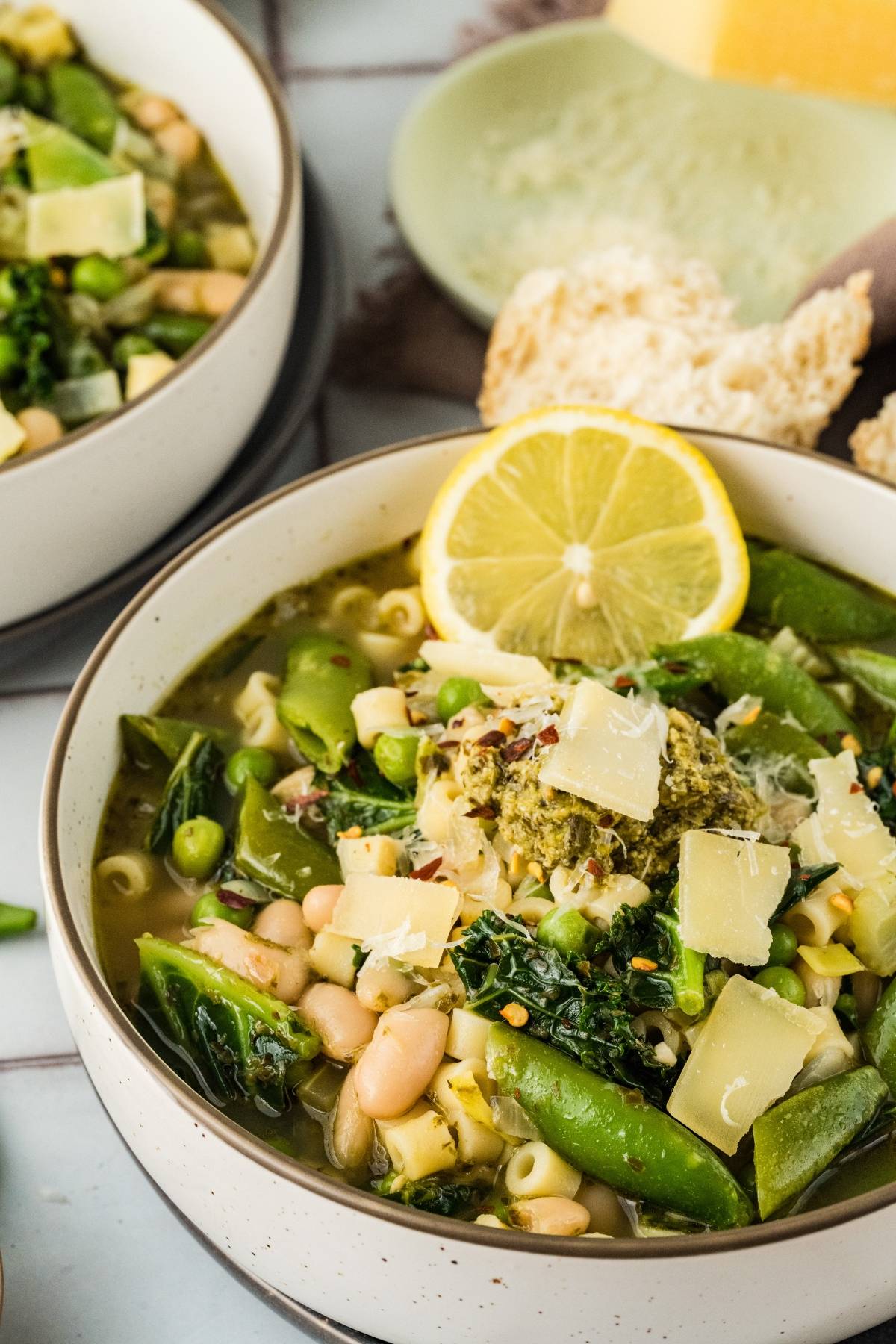 A bowl of vegetable soup with pasta, beans, greens, shaved cheese, and a lemon slice, served with bread.