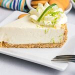 A slice of no bake key lime cheesecake with whipped cream and lime zest on a white plate, surrounded by fresh limes and cookies.