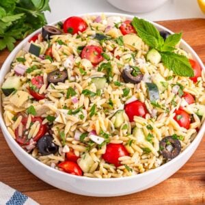 A vibrant bowl of orzo salad with tomatoes, cucumbers, olives, fresh herbs, and lemon sits on a wooden board.
