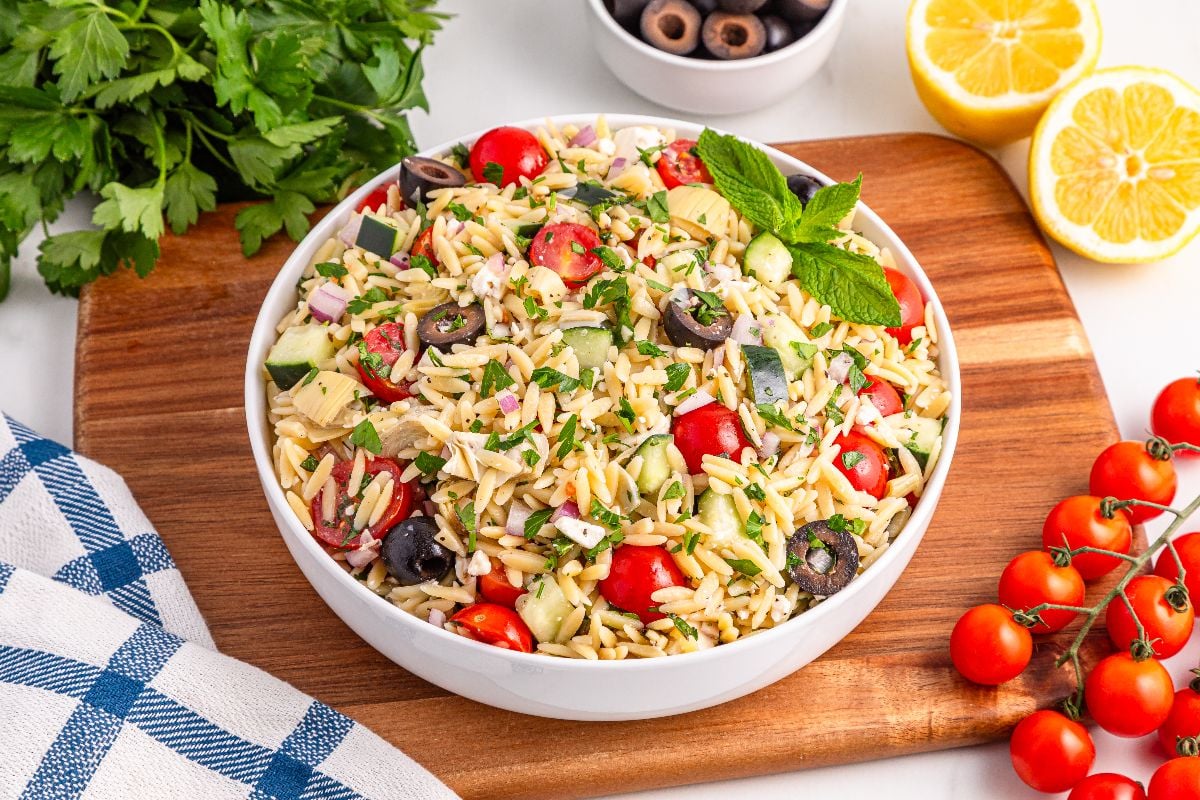 A bowl of orzo salad with tomatoes, cucumbers, olives, and herbs sits on a wooden board surrounded by fresh ingredients.