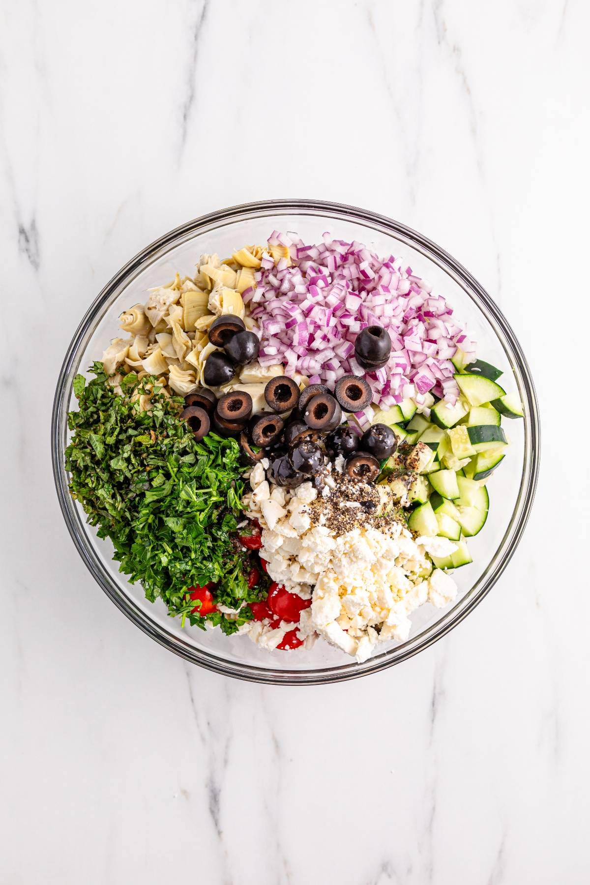 A glass bowl filled with chopped vegetables, olives, herbs, and feta cheese arranged in sections.