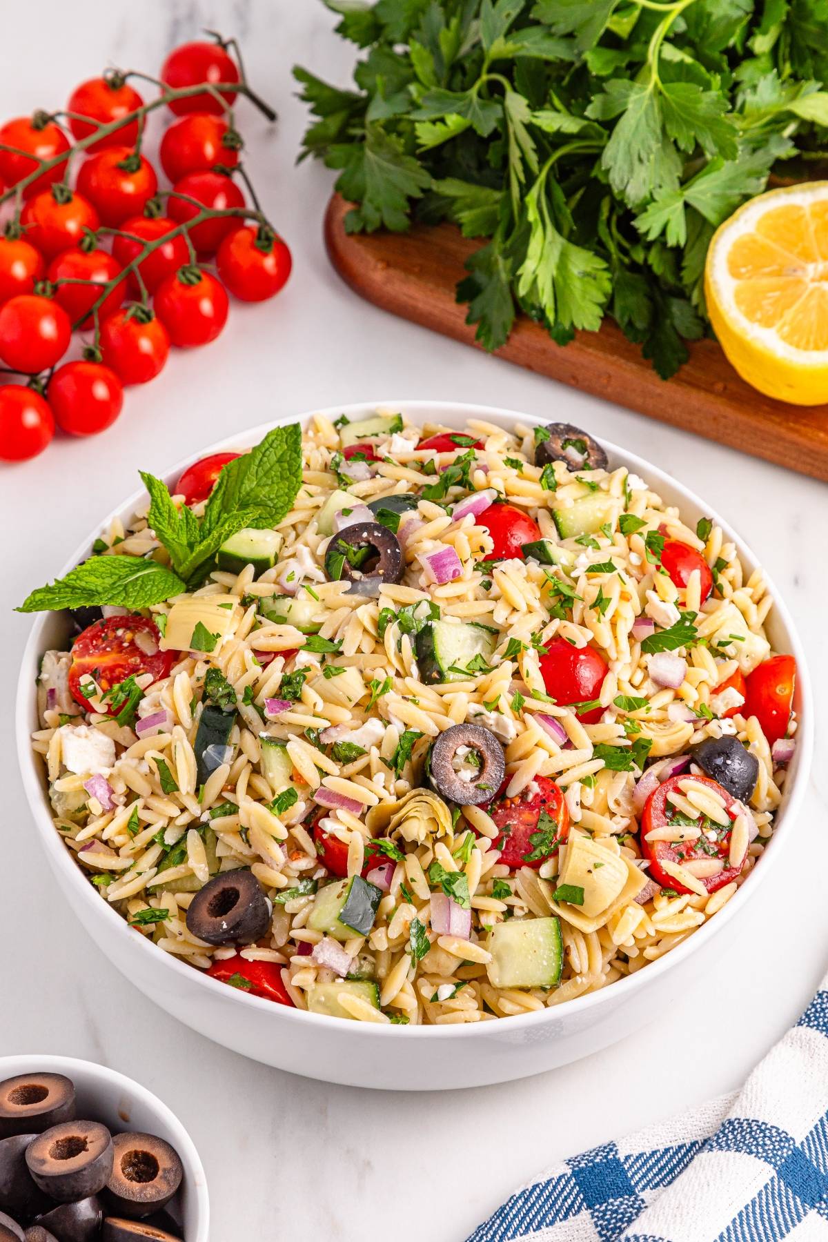 Bowl of orzo pasta salad with olives, tomatoes, cucumbers, and herbs, surrounded by fresh veggies and lemon.
