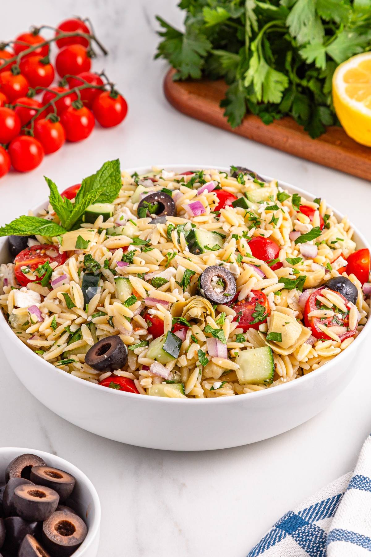 A bowl of orzo pasta salad with olives, tomatoes, cucumbers, and herbs sits on a marble countertop.