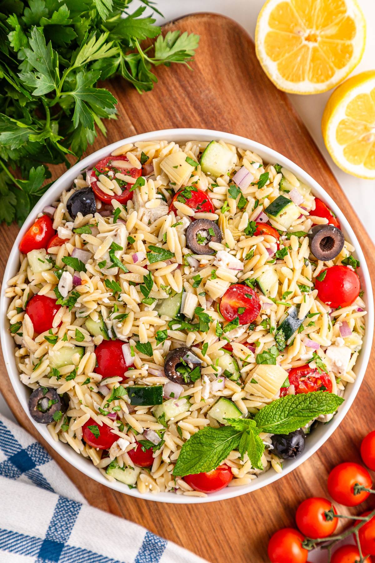 A bowl of orzo salad with tomatoes, cucumbers, olives, herbs, and lemon on a wooden board.