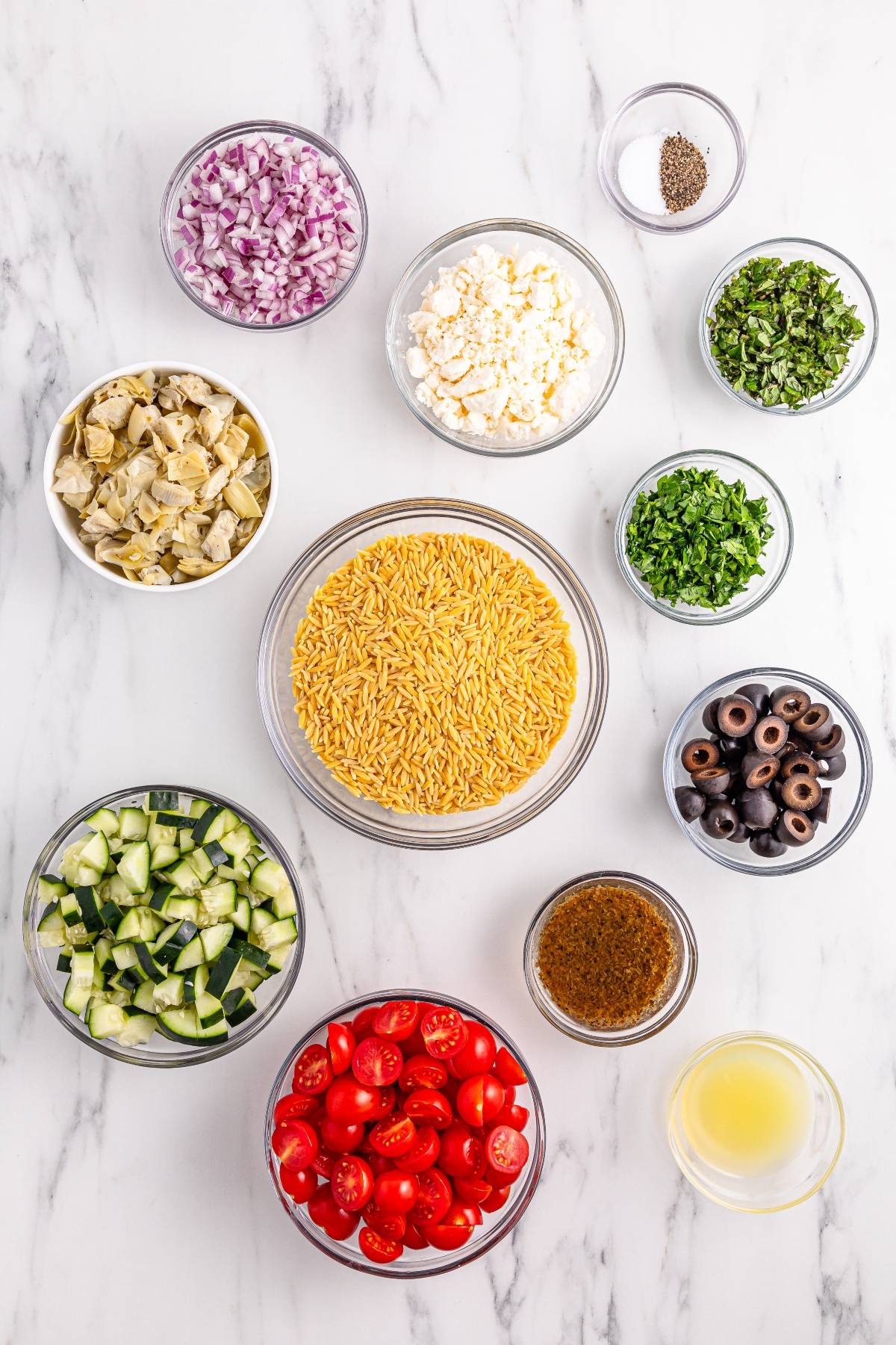 Bowls of chopped red onion, artichoke, olives, tomatoes, cucumber, herbs, feta, orzo pasta, and dressings on marble.