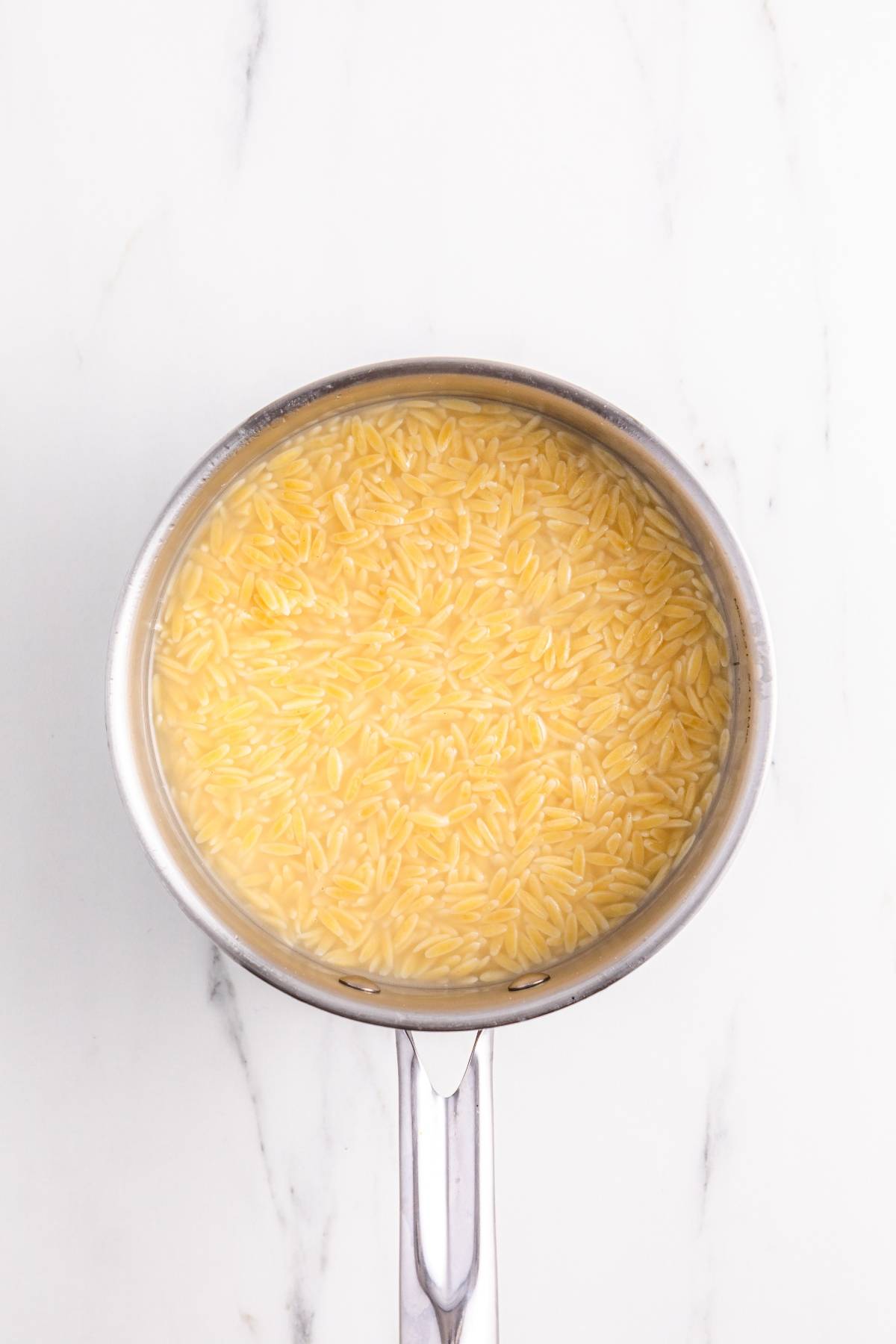 A pot of uncooked orzo pasta soaking in water on a white marble surface.