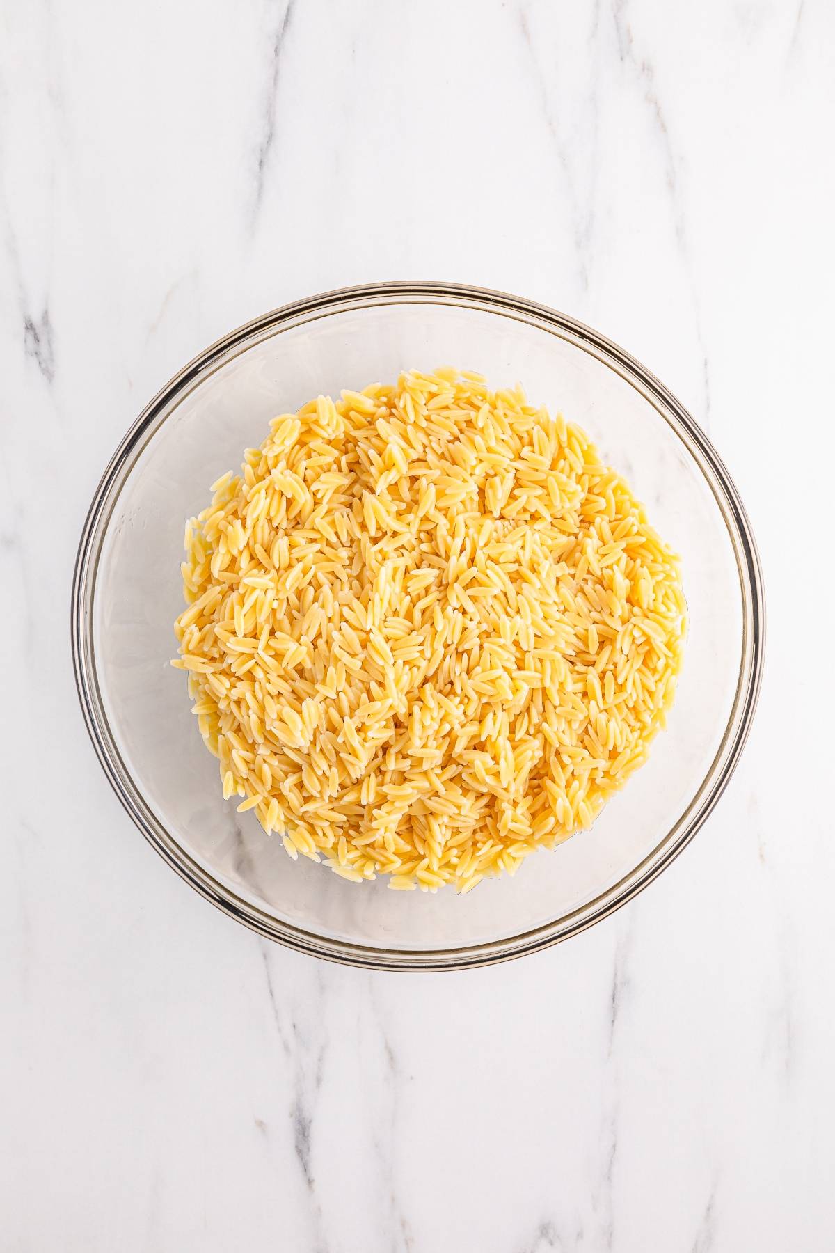 A glass bowl filled with cooked orzo pasta on a white marble surface.
