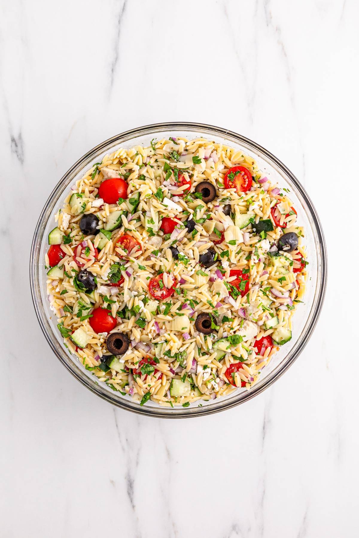 A glass bowl filled with orzo salad, cherry tomatoes, olives, cucumbers, and herbs on a marble surface.