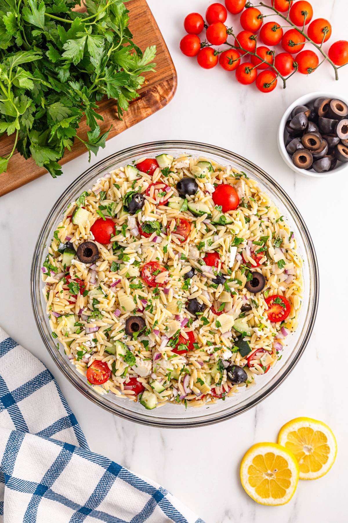 A bowl of orzo pasta salad with tomatoes, olives, cucumbers, and herbs, surrounded by fresh ingredients.