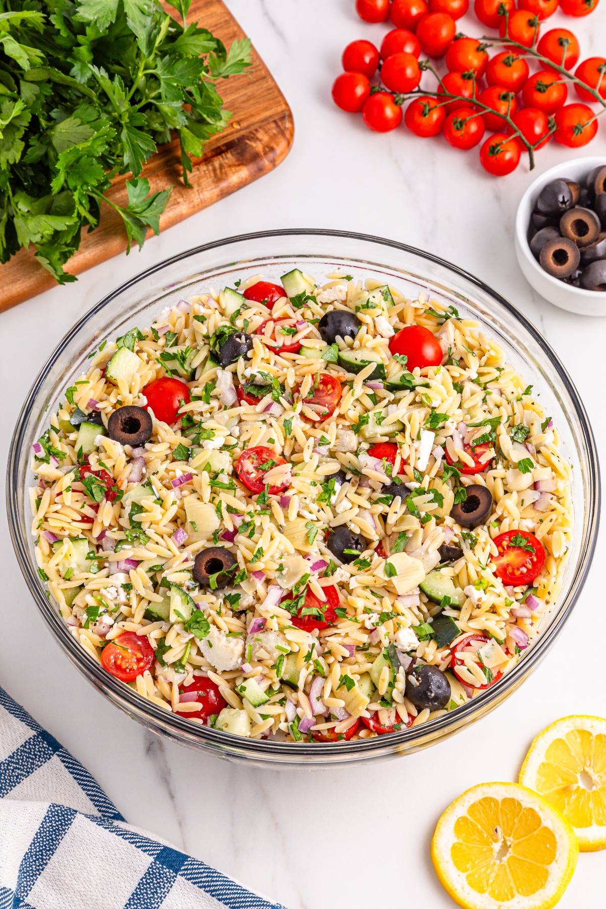 A glass bowl filled with orzo pasta salad with tomatoes, olives, cucumber, and parsley on a marble surface.