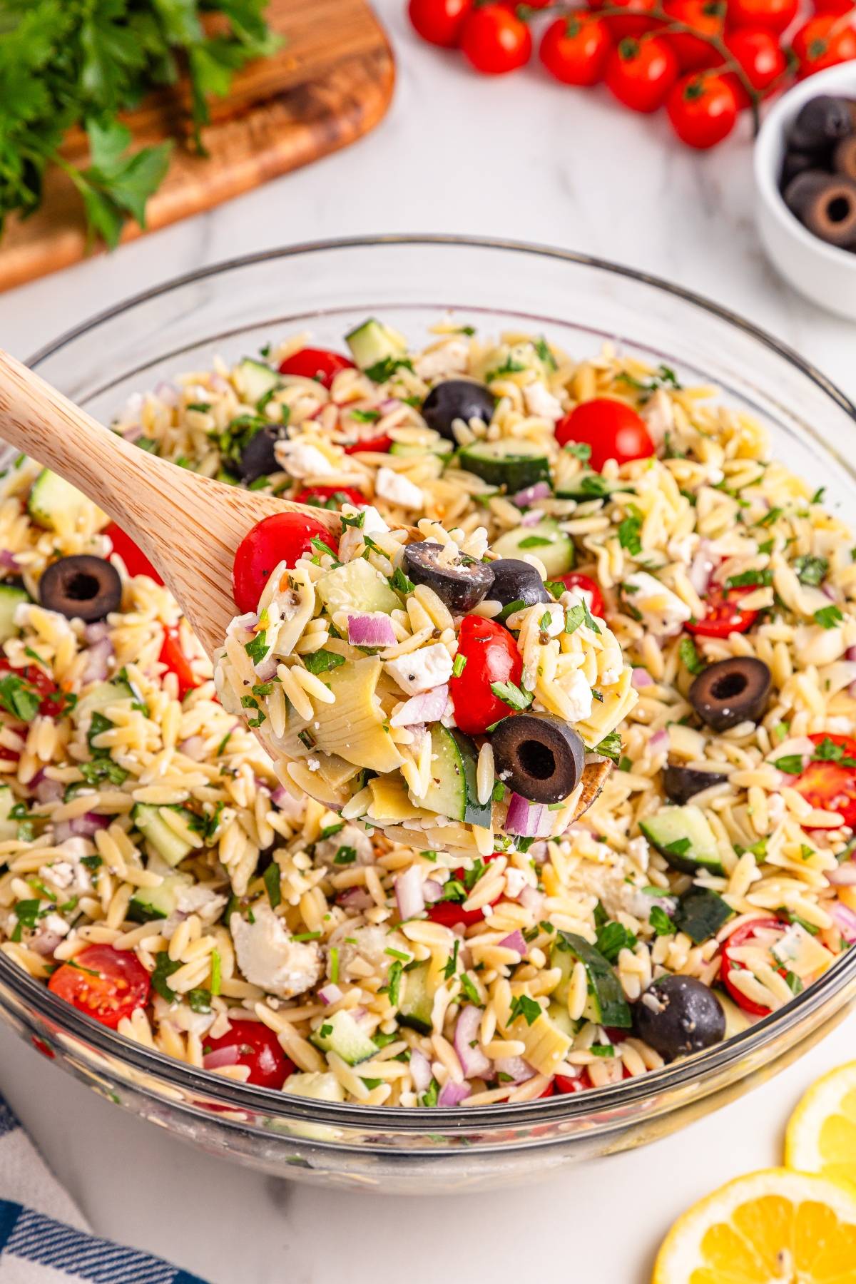 A bowl of orzo pasta salad with tomatoes, olives, cucumbers, onions, and herbs, scooped with a wooden spoon.