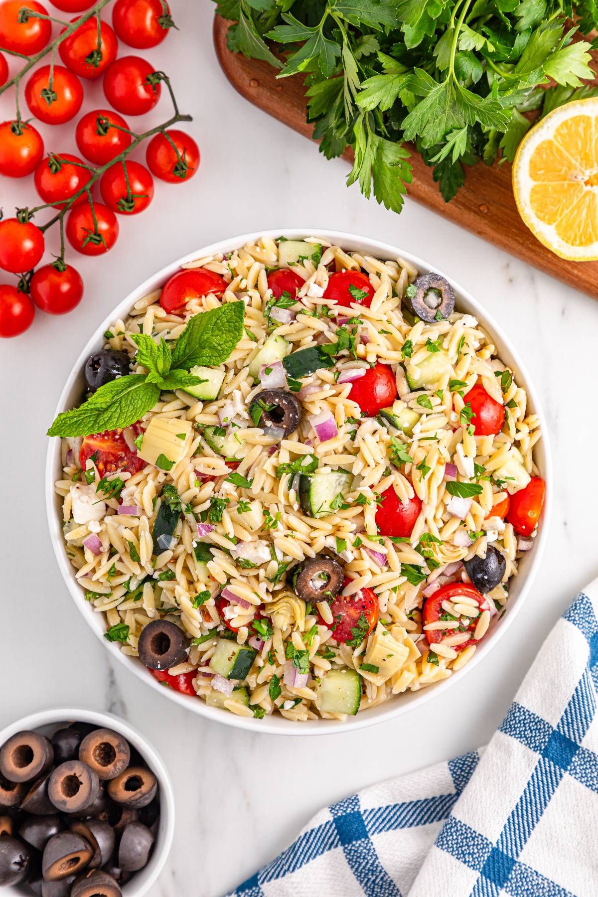 A bowl of orzo pasta salad with tomatoes, olives, cucumber, artichoke, and herbs on a table with fresh ingredients.