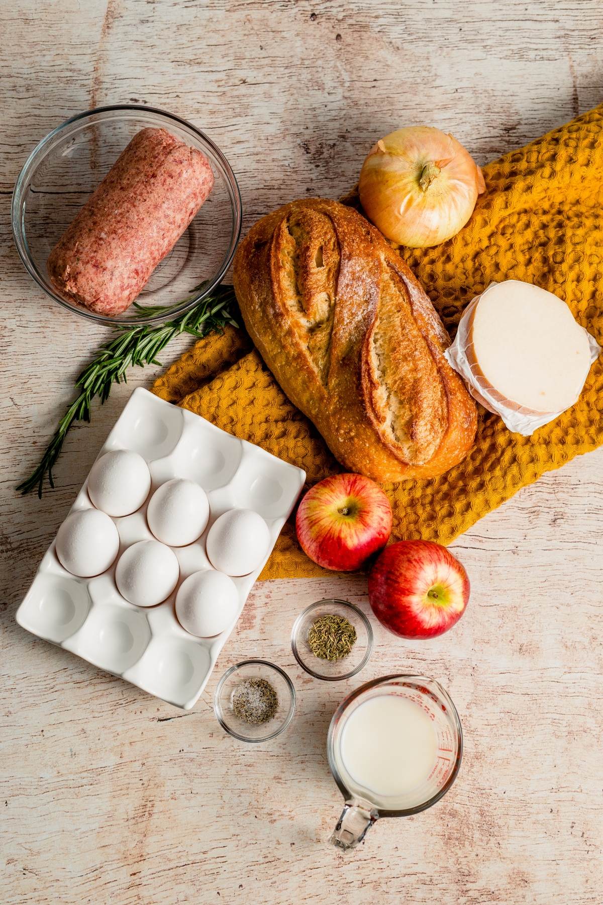 Ingredients on a table: eggs, bread, apples, onions, sausage, cheese, herbs, and milk on a yellow cloth—perfect for preparing a delicious Sausage Apple Breakfast Casserole.