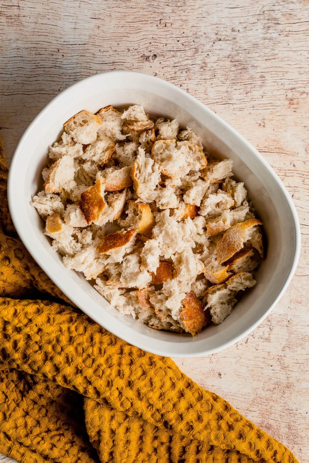 A white dish filled with torn bread pieces sits on a textured mustard-colored cloth, perfect for assembling a Sausage Apple Breakfast Casserole that’s ideal for a cozy Thanksgiving or Christmas breakfast.